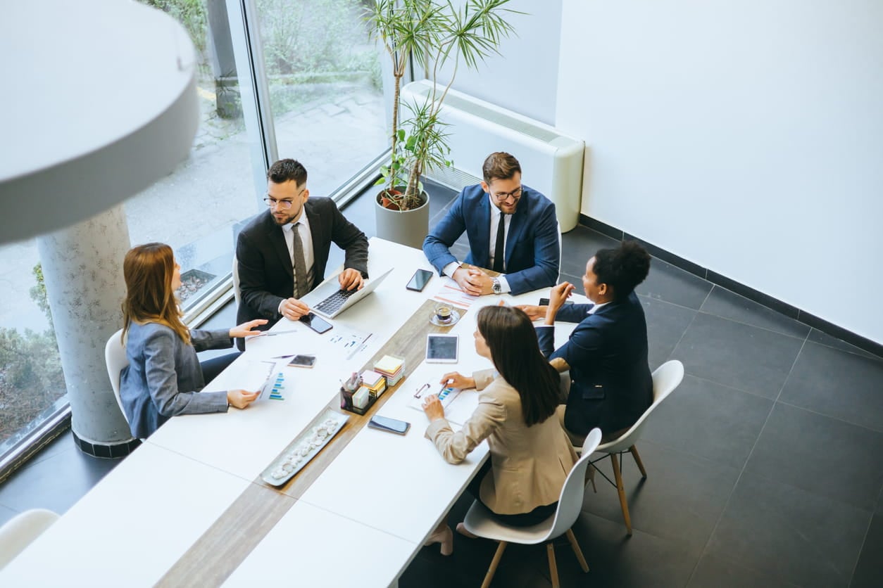 Five suited professionals meet around a white conference table with laptops and papers, discussing in a bright office.